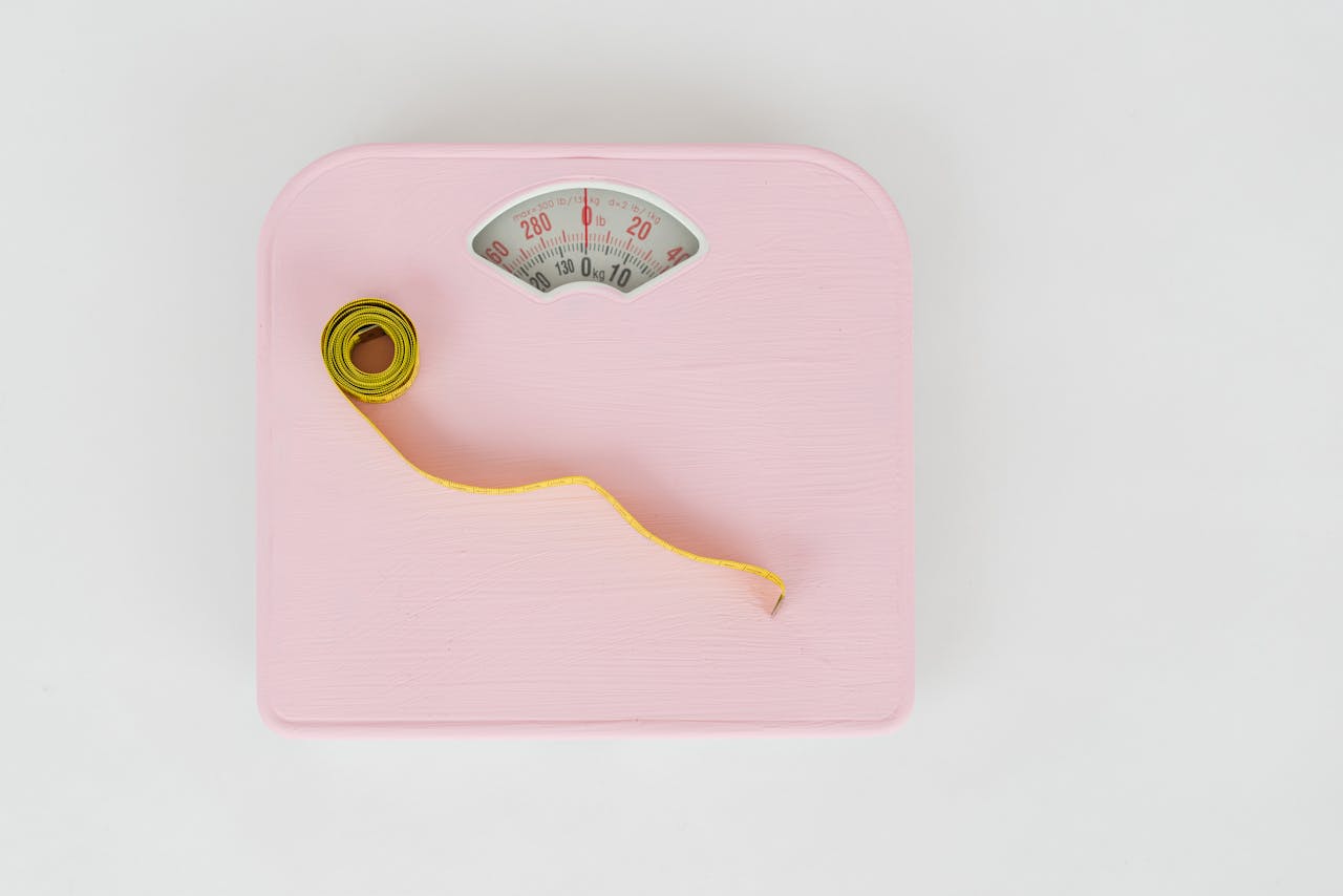 who-we-are A pink weighing scale paired with a yellow measuring tape on a white backdrop.